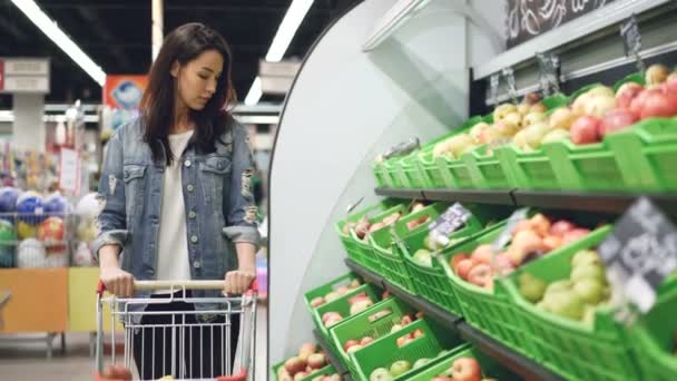 Charming girl in casual clothing is walking along fruit row moving shopping trolley and looking at organic fruit with smile. Healthy lifestyle and supermarket concept. - Video
