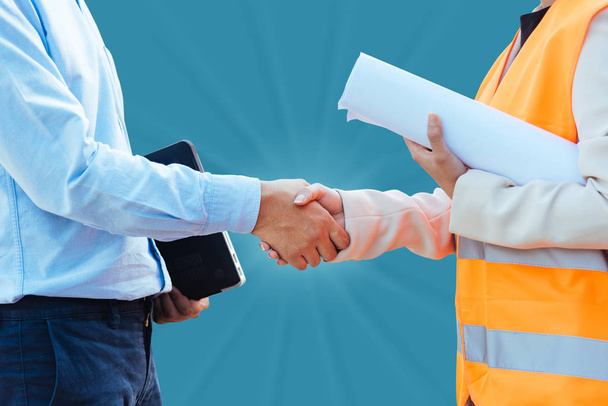 Close up Asian man civil engineer holding laptop and Asian woman architect holding drawing blueprint shaking hands isolated on blue background. - Фото, изображение