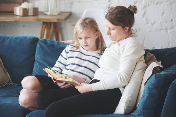 daughter reading book while sitting with mom on couch in apartment  - Photo, image