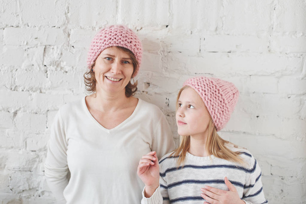Mother and daughter in matching pink knitted hats posing on background of white brick wall - Foto, immagini