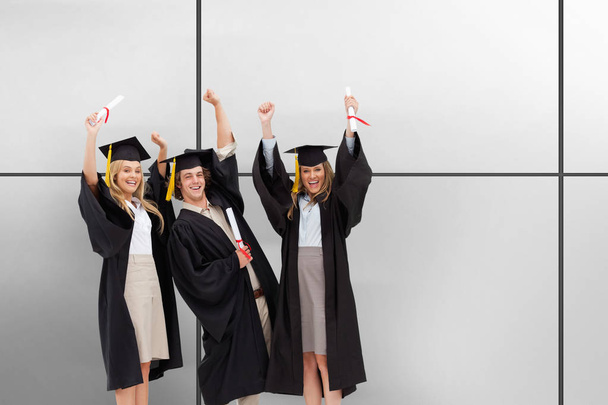 Three students in graduate robe raising their arms against white tiling - Foto, Bild