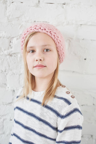 Portrait of blonde girl in knitted pink hat looking at camera on white brick wall background  - Φωτογραφία, εικόνα