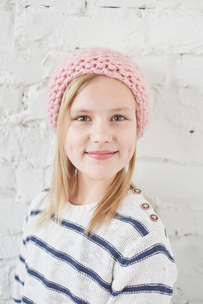Portrait of blonde girl in knitted pink hat looking at camera on white brick wall background  - Photo, image