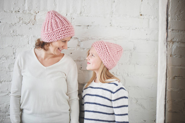 Mother and daughter in matching pink knitted hats posing on background of white brick wall - Zdjęcie, obraz