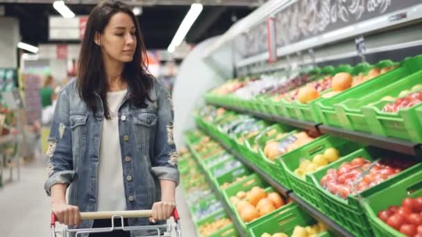 Pretty young woman is walking along fruit and vegetable row pushing shopping trolley and looking at organic food with smile. Healthy lifestyle and supermarket concept. - Imágenes, Vídeo