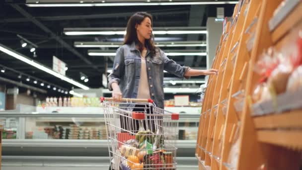 Smiling girl is buying fresh bread in supermarket smelling it then putting in cart with other products. Shopping for food, cheerful people and happy customers concept. - Video