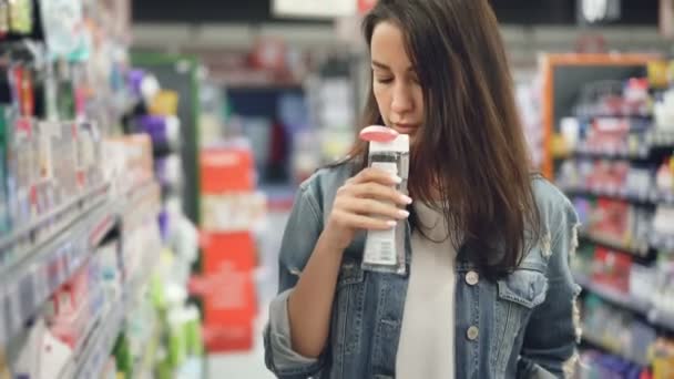 Young woman is buying cosmetics in supermarket, she is opening bottle with liquid and smelling it then smiling and putting in trolley. People and goods concept. - Filmagem, Vídeo