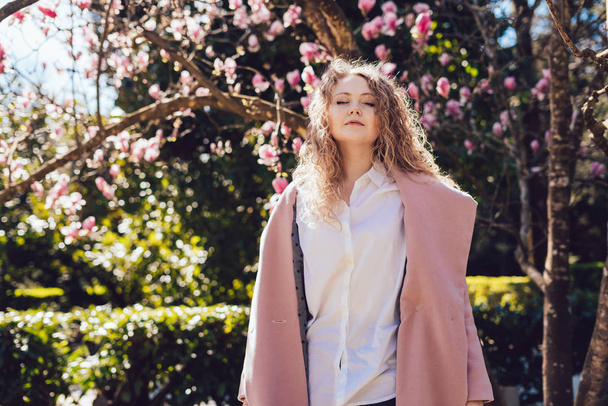 elegant curly girl in a trendy pink coat posing in the park in the sun, enjoying the spring - Фото, изображение