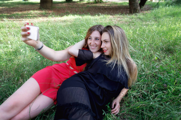 Duas garotas esticadas na grama tiram uma selfie juntas e sorriem. No fundo o verde no parque ao ar livre
. - Foto, Imagem