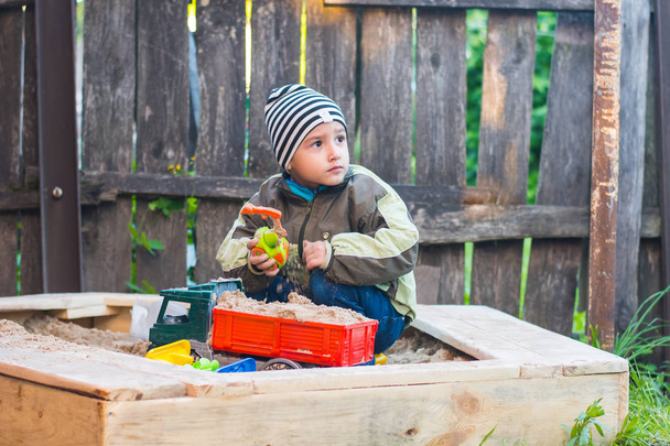 boy in a striped hat playing in a sandbox in the yard - Photo, Image