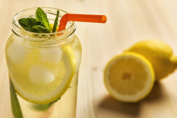 Close-up of the top of a glass jar with lemonade on a wooden table and some unfocused lemons in the background. The jug contains water, slices of lemon, ice, mint leaves and a cane to drink - Zdjęcie, obraz