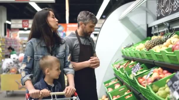 Sociable handsome shop assistant is selling fresh fruit to attractive young woman with child, man is pointing at bright boxes with products and speaking. - Materiał filmowy, wideo