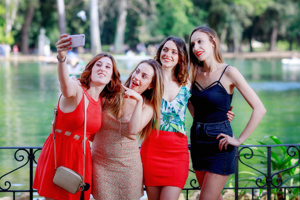 Τέσσερα κορίτσια, φίλους, χαμόγελο και πάρτε μια selfie μαζί. Η ξανθιά κοπέλα εγείρει αγκαλιά ως σήμα της νίκης. Στο παρασκήνιο η πράσινη λίμνη στο δημόσιο πάρκο. - Φωτογραφία, εικόνα