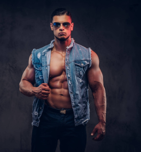 Sexy naked fashionable athletic man in a denim vest and sunglasses poses in a studio. - Photo, Image