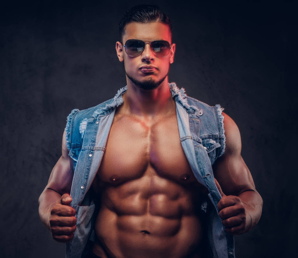 Sexy naked fashionable athletic man in a denim vest and sunglasses poses in a studio. - Fotografie, Obrázek