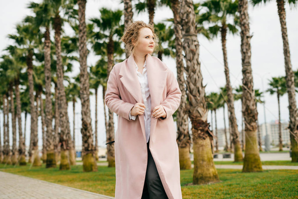 a nice stylish girl in a fashionable pink coat walks the park next to tall green palms, enjoying spring and warmth - Foto, afbeelding
