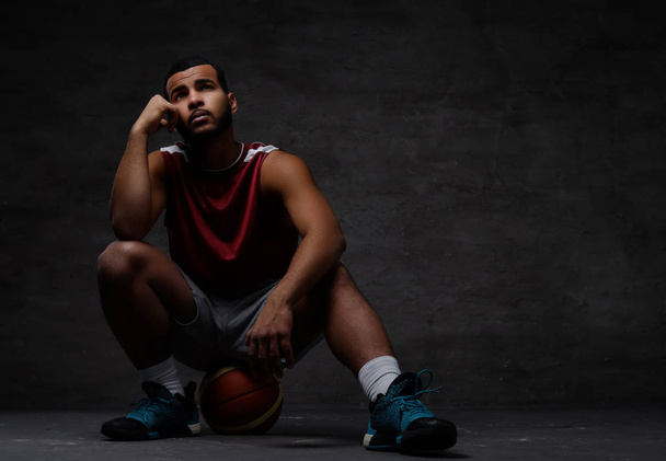 Zamyšlený mladé afroamerické basketbalový hráč v oblečení sedí na kouli nad tmavým pozadím. - Fotografie, Obrázek