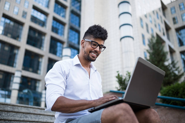 Young handsome Afro American man sitting in front of huge modern business building, holding business card and doing some work on laptop.  - Foto, Imagen