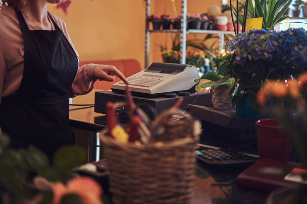 Charming redhead female florist wearing uniform works with cash register in flower shop. - Photo, Image