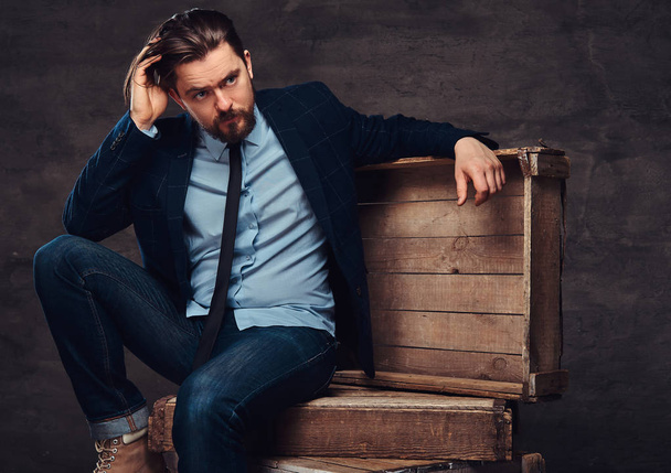 Portrait of a middle age businessman with stylish hair and beard dressed in jeans, jacket and tie, sitting on wooden boxes in a studio. - Photo, Image