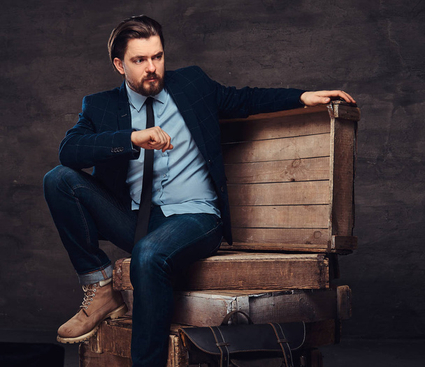 Portrait d'un homme d'affaires d'âge moyen avec des cheveux et une barbe élégants vêtus de jeans, veste et cravate, assis sur des boîtes en bois dans un studio
. - Photo, image