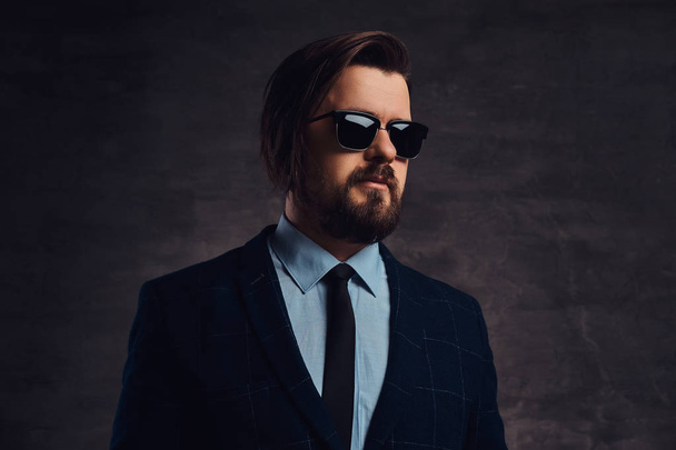 Close-up portrait of a handsome fashionable middle-aged man with beard and hairstyle dressed in an elegant formal suit and sunglasses on a textured dark background in studio. - Photo, Image