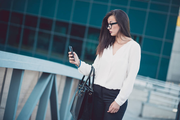 Krásná bruneta žena na sobě bílou košili textilie na chytrém telefonu chůzi na ulici v zamračený den. - Fotografie, Obrázek