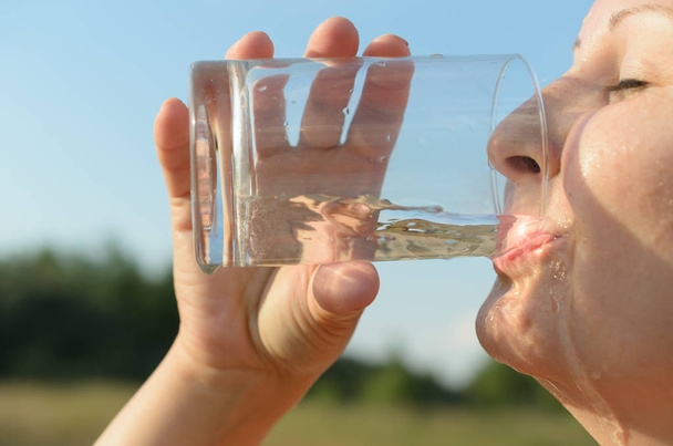 eine einfache europäische junge Frau trinkt vor blauem Himmel Wasser aus einem Glasbecher. - Foto, Bild