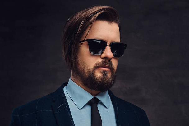 Close-up portrait of a handsome fashionable middle-aged man with beard and hairstyle dressed in an elegant formal suit and sunglasses. Isolated on a textured dark background in studio. - Photo, Image