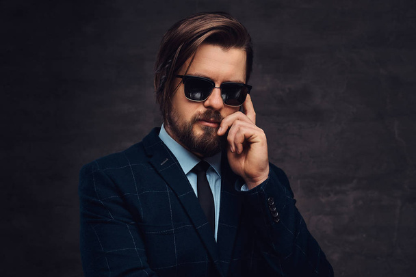 Close-up portrait of a handsome fashionable middle-aged man with beard and hairstyle dressed in an elegant formal suit and sunglasses. Isolated on a textured dark background in studio. - Foto, Imagen