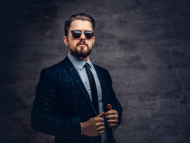 Confiado guapo hombre de mediana edad de moda con barba y peinado vestido con un elegante traje formal y gafas de sol sobre un fondo oscuro en el estudio
. - Foto, imagen