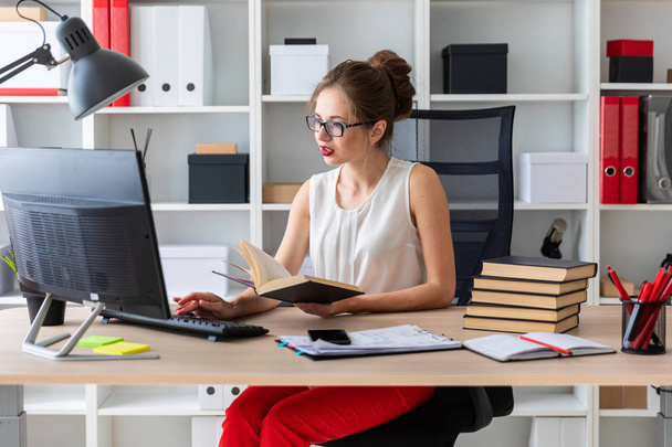 Una joven se sienta en una mesa de computadora y sostiene un libro abierto en sus manos. - Foto, Imagen