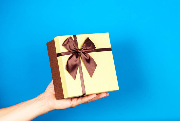 Human hand holding brown ribbon wrapped holiday surprise gift or present box package on blue background - Photo, Image