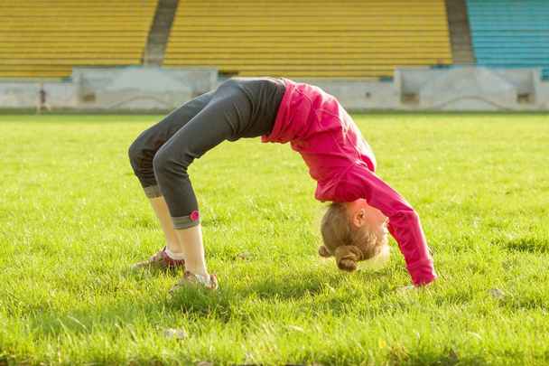 Спортивна маленька дівчинка стоїть догори ногами на зеленій траві на стадіоні. Концепція здорового способу життя
 - Фото, зображення