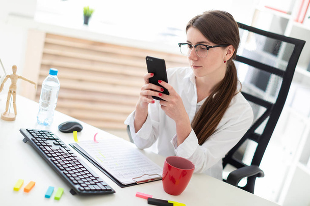 Una joven está sentada en una mesa en la oficina, mirando el teléfono y al lado hay una taza
. - Foto, imagen