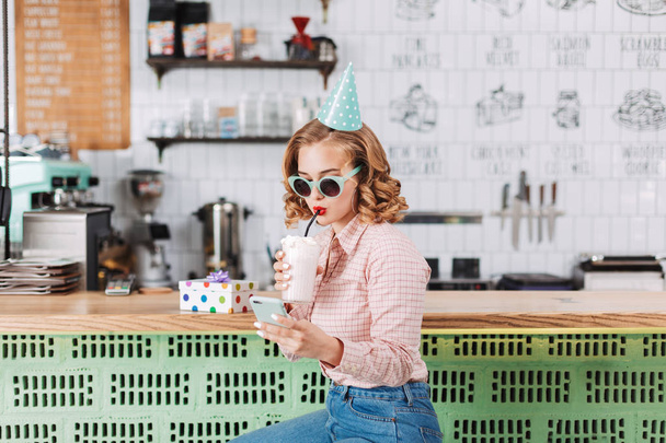 Güzel kız güneş gözlüğü ve barda oturan Doğum günü şapkası sayaç ve kafede onun cep telefonu kullanırken milkshake içme - Fotoğraf, Görsel