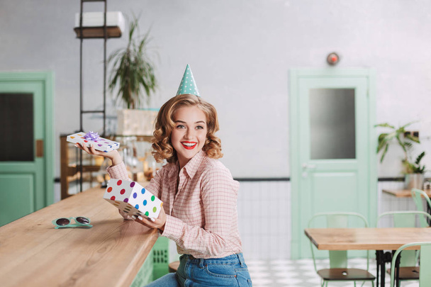 Молода усміхнена леді в шапці на день народження сидить за барною стійкою з відкритою подарунковою коробкою в руках і щасливо виглядає в кав'ярні
 - Фото, зображення