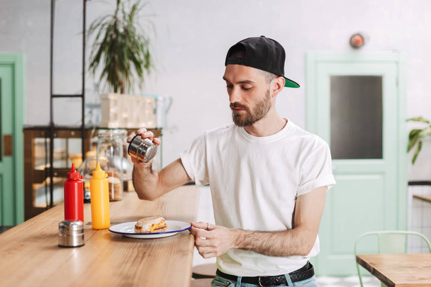 Jeune homme réfléchi en bonnet noir assis au comptoir du bar et tenant du sel tout en mangeant un hamburger dans un café
 - Photo, image
