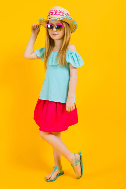 Girl with red hair on a yellow background. A charming girl in bright summer clothes holds her hand for her hat. - Foto, Imagen