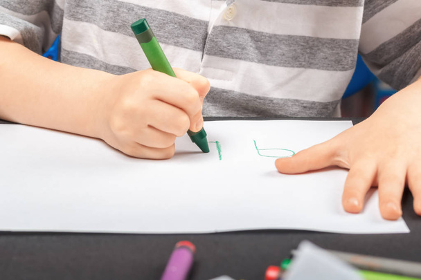 Drawing on a white sheet of paper with colored wax pencils. The concept of childrens creativity and hobbies. A child draws a green pencil on white paper. Baby hands closeup - Фото, изображение
