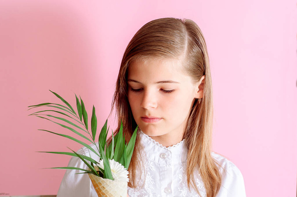 girl holding ice cream cone with white flowers daisy and a green branch of a fern and looking at camera on pink background - Photo, Image