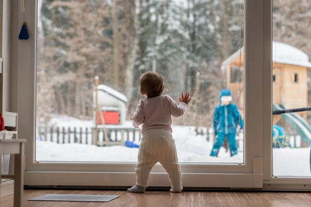 Little baby standing at a large view window watching a sibling outdoors playing in thick winter snow in the garden with forest backdrop. - Фото, изображение