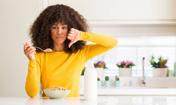 Afrikai-amerikai nő eszik gabona és tej otthon, haragos arc, negatív előjellel mutatja ellenszenv a hüvelykujját lefelé, elutasítás koncepció - Fotó, kép