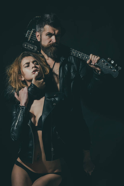 Rock and roll concept. Couple in love cuddling with guitar, black background. Guitarist with beard and sexy girl in bra enjoy rock and roll lifestyle. Couple sexy and brutal posing with guitar - Photo, Image