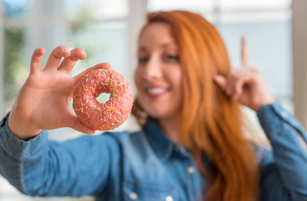 Redhead woman holding donut at home surprised with an idea or question pointing finger with happy face, number one - Фото, изображение