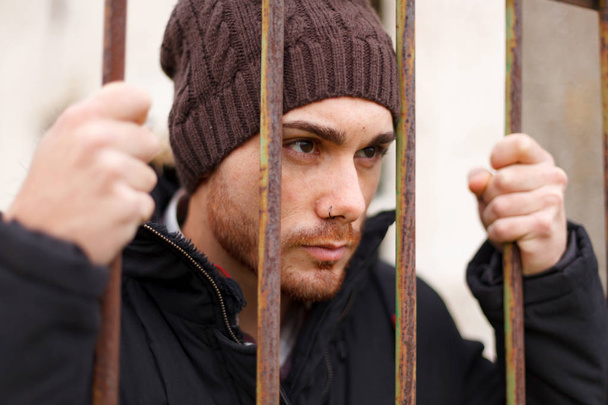 attractive young man in coat and knitted cap posing behind the bars  - Photo, image