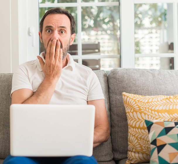 Мужчина средних лет, использующий ноутбук на покрывала дивана рот с рукой шокирован стыдом за ошибку, выражение страха, боится в тишине, тайная концепция
 - Фото, изображение