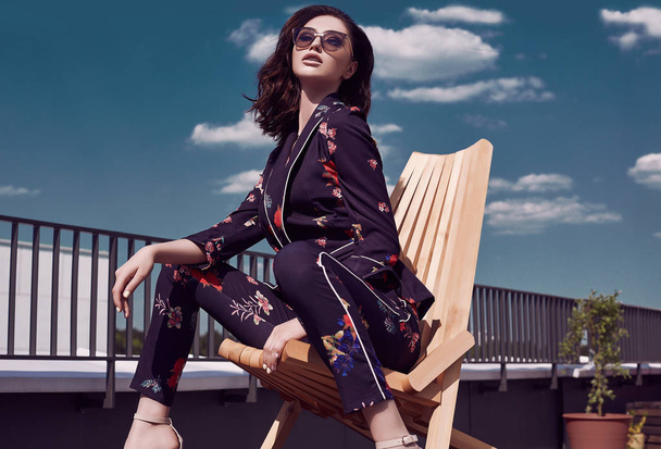 Portrait of a gorgeous bright brunette woman in vivid dress sitting in a wooden chair on the roof of a building - Photo, Image