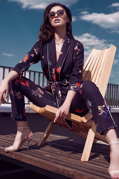 Portrait of a gorgeous bright brunette woman in vivid dress sitting in a wooden chair on the roof of a building - Photo, Image