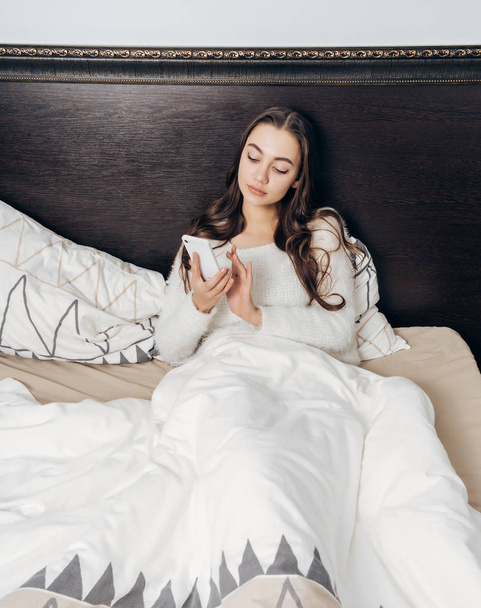 young sleepy girl in white pajamas lies in bed, puts an alarm clock on her smartphone - Fotó, kép